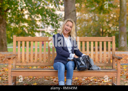 Attraktive blonde Frau mit einem Lächeln erfreut von Stöbern in ihrer Handtasche, als sie auf einer hölzernen Parkbank sitzt Stockfoto