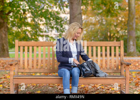 Attraktive blonde Frau mit einem Lächeln erfreut von Stöbern in ihrer Handtasche, als sie auf einer hölzernen Parkbank sitzt Stockfoto