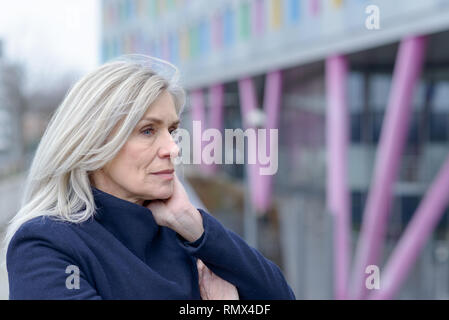 Nachdenkliche Frau, die in die Ferne starrt, als sie auf das Geländer stützt sich auf einem städtischen Gehweg ihr Kinn auf ihre Hände in der Nähe zu Profil p Stockfoto