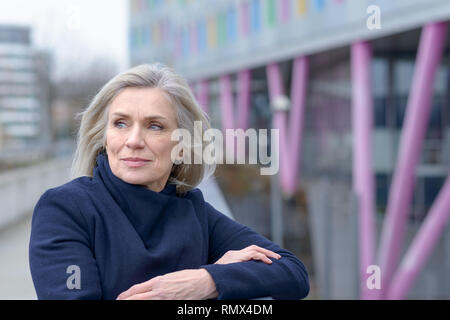 Nachdenklich reife attraktive blonde Frau Blick zurück über die Schulter zu, wie sie sich in Anlehnung an den Rand Geländer einer städtischen Außen walkwa Stockfoto