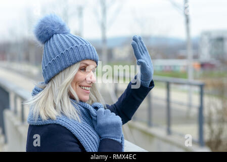 Gerne blonde Frau in warmen Winter woollies winkt ein Gruß da steht sie lächelnd auf einem Gehweg im Freien in der Stadt Stockfoto