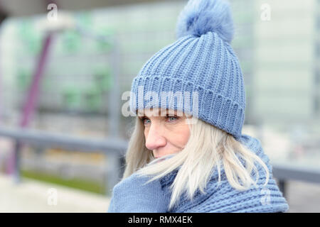 Attraktive blonde Frau in einem trendigen Blau winter Outfit kuschelte sich in einen warmen Schal hält Ihre behandschuhten Hände zu ihr Kinn, als sie steht draußen warten Stockfoto