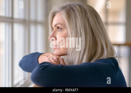 Attraktive blonde Frau beobachten, die durch ein Fenster mit einem ernsten Ausdruck ihr Kinn auf ihre Hände, als sie auf einer hölzernen Geländer lehnt. Stockfoto