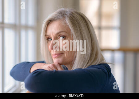 Attraktive blonde Frau beobachten, die durch ein Fenster mit einem ernsten Ausdruck ihr Kinn auf ihre Hände, als sie blickt in die Kamera Stockfoto