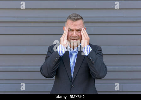 Betont, Geschäftsmann, oder leiden unter Kopfschmerzen, stehend mit den Händen die Schläfen Grimassen gegen einen grauen Außenwand Stockfoto