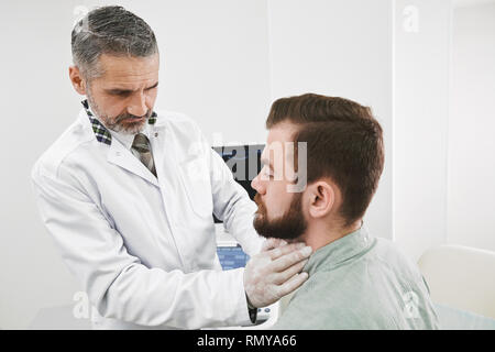 Professionelle Therapeuten abtasten, Kontrolle Lymphknoten am Hals des Patienten. Der bärtige Mann sitzt auf Rücksprache mit Arzt in der Klinik. Schwere Arzt tragen in der medizinischen einheitliche und Handschuhe. Stockfoto