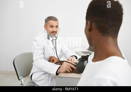 Arzt mit Blutdruckmessgerät für die Messung von Blutdruck des Patienten. Afrikanische Mann sitzt auf Beratung, die Hand auf den Tisch. Arzt tragen in der medizinischen weißes Kleid. Stockfoto