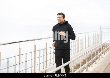 Bild von zufriedenen Sportler 30 s in schwarz Sportswear entlang Pier am Meer Stockfoto