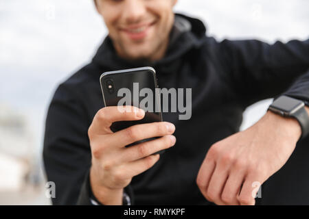Bild von Reifen Sportsman 30 s in schwarz Sportbekleidung mit Smartphone beim Sitzen auf der Promenade am Meer Stockfoto