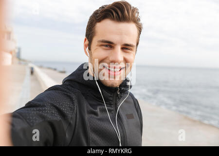 Bild von lächelnden Sportler 30 s in schwarz Sportswear- und Ohrhörer unter selfie Foto auf Handy beim Spaziergang am Meer Stockfoto