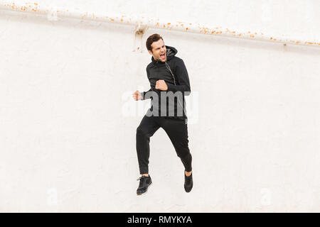 Bild der europäischen Mann 30 s in schwarz Sportswear- und Ohrhörer entlang der weißen Wand laufen Outdoor Stockfoto