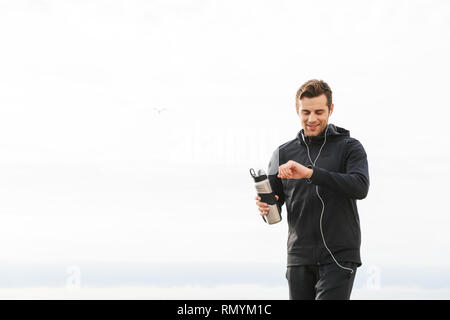 Bild von zufriedenen Sportler 30 s in schwarz Sportswear- und Ohrhörer aus und läuft durch Seaside Stockfoto