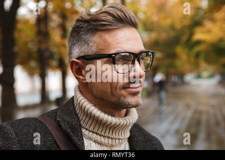 Foto von bärtigen Mann 30 s Tragen von Brillen an Kamera schaut beim Gehen im Freien durch Herbst Park Stockfoto