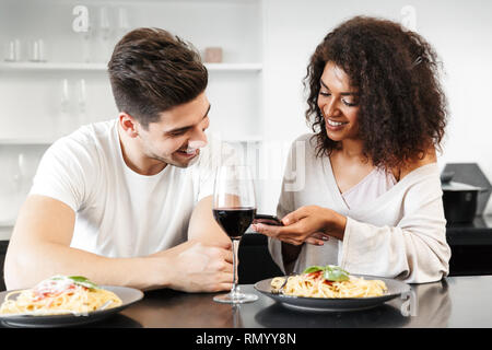 Schönen jungen multiethnischen Paar für ein romantisches Abendessen zu Hause, trinken Rotwein und essen Pasta, mit Handy Stockfoto