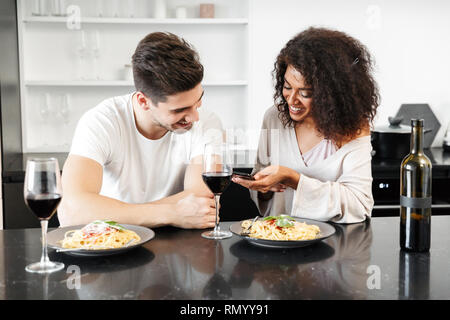Schönen jungen multiethnischen Paar für ein romantisches Abendessen zu Hause, trinken Rotwein und essen Pasta, mit Handy Stockfoto