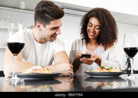 Schönen jungen multiethnischen Paar für ein romantisches Abendessen zu Hause, trinken Rotwein und essen Pasta, mit Handy Stockfoto
