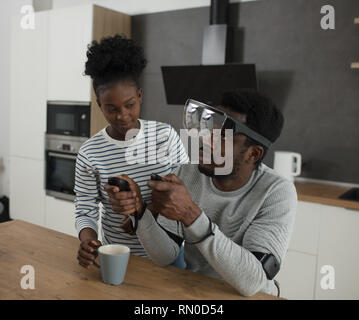 Afrikanischer mann Vornehmen der Einstellungen der virtuellen Realität Konsole mit Controller, während seine Frau ihm die Vorbereitung am Morgen Kaffee in der modernen Küche Stockfoto