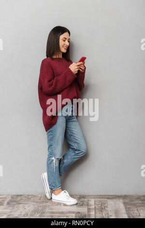 Bild der hübschen jungen Frau in Burgund Pullover mit Handy über graue Wand Hintergrund isoliert gekleidet. Stockfoto
