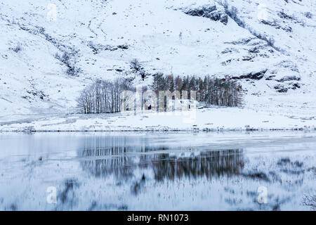 Snowy Reflections mit Bäumen und Cottage im Loch Achtriochtan, Glencoe, Scottish Highlands, Schottland Großbritannien im Januar Stockfoto