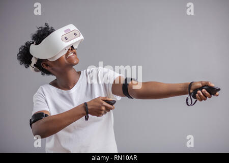 Schöne junge afrikanische Frau im weißen T-Shirt tragen weiße virtual reality Headset und hält Joysticks, posing ar Kamera über grauen Hintergrund. Stockfoto