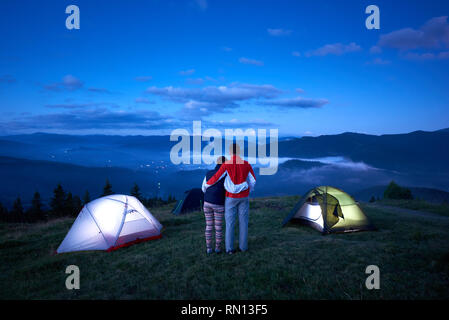 Paar steht in der Nähe von Camping mit dem Rücken zur Kamera und umarmen einander genießen Sie den Sonnenaufgang in den Bergen. Leichter Dunst über das Tal zwischen den Bergen, durch die die Lichter der Stadt sichtbar sind Stockfoto
