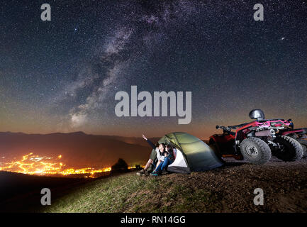 Junges Paar, Mann und Frau sitzen zusammen im Zelt auf dem Gipfel des Berges mit ATV Quad Motorrad. Mann an schöne Nacht Himmel voller Sterne zeigen, Milchstraße, leuchtende Stadt auf Hintergrund Stockfoto