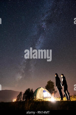 Silhouette von romantisches Paar Wanderer mit einer Rast am Lagerfeuer neben Camp und touristische Zelt unter erstaunlichen Nacht Himmel voller Sterne und Milchstraße. Auf Hintergrund Sternenhimmel, Berge und leuchtende Stadt Stockfoto