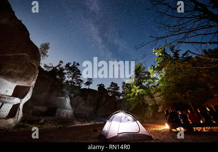 Ansicht der Rückseite des Freunde Touristen zusammen sitzt auf der Bank neben dem Camp, Bonfire und leuchtende Zelt in der Nacht. Auf Hintergrund schöne Nacht sternenklaren Himmel voller Sterne und die Milchstraße, Felsen, Bäume Stockfoto