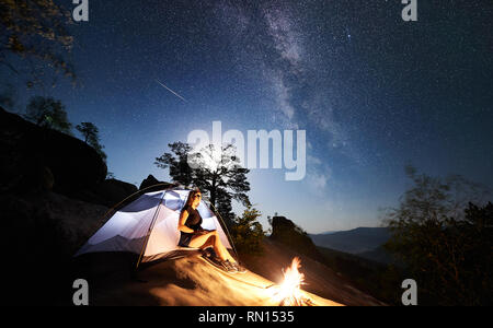 Wanderer Frau ruht auf Rocky Mountain Top neben Camp, Bonfire und touristische Hütte im Sommer Nacht und genießen Sie zauberhafte Sicht der Nacht Himmel voller Sterne und Milchstraße. Auf Hintergrund Sternenhimmel und Bäume Stockfoto