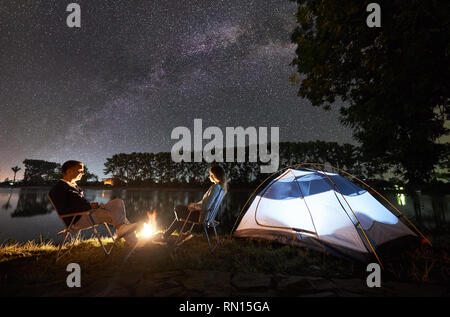 Nacht Camping auf See. Mann und Frau Freunde sitzen auf Stühlen in der Nähe von Lagerfeuer und beleuchteten Zelt, genießen Sie unglaubliche Aussicht auf Abend Himmel voller Sterne und die Milchstraße, die Lichter der Stadt im Hintergrund. Stockfoto