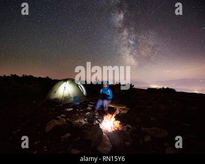 Weibliche Reisemobile mit einem Rest an Sommer Nacht Camping in den Bergen neben Lagerfeuer und beleuchtete touristische Hütte. Junge Frau Backpacker genießen Sicht der Nacht Himmel voller Sterne und Milchstraße. Stockfoto