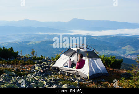 Camping auf der Spitze des Berges, auf hellen Sommermorgen. Rückansicht der Frau in der Eingang der touristischen Zelt sitzen. Auf nebligen Berge im Hintergrund. Tourismus Abenteuerurlaub active lifestyle Konzept Stockfoto