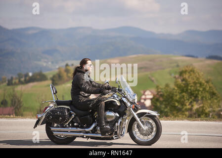 Seitenansicht des bärtigen langhaarigen Motorradfahrer mit Sonnenbrille und schwarzem Leder kleidung Reiten cruiser Motorrad auf schmalen asphaltierten Weg an sonnigen Herbsttag. Stockfoto