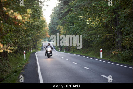 Motorbiker in Helm und schwarzem Leder Bekleidung, moderne leistungsfähige Motorrad entlang der asphaltierten Straße Wicklung unter hohen grünen Bäumen, an sonnigen Herbsttag. Stockfoto