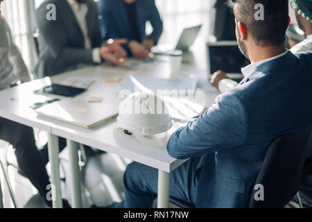 7/8-Ansicht der männlichen Ingenieure und Konstrukteure in der Sitzung an Chief Executive Office teilnehmenden, Sitzen am Tisch mit Laptop und harten Hut auf. Stockfoto