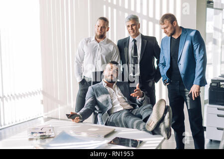 Lächelnd entspannte Chef im Anzug in seinem Büro sitzen an Kamera mit ausgereiften Assistant und zwei Bodyguards im Hintergrund suchen, Stockfoto