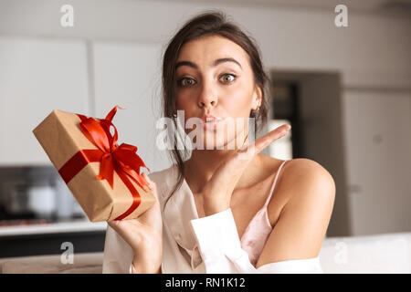 Attraktive junge Frau auf einer Couch zu Hause sitzen, halten eine Geschenkverpackung Stockfoto