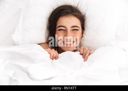 Bild von der Oberseite der friedlichen Frau 20s mit dem dunklen Haar lachend, während im Bett unter weißen Leinen liegend Stockfoto