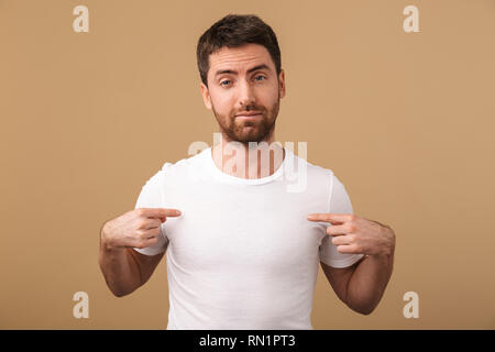 Porträt eines umgekippten junger Mann beiläufig stehend über beige isoliert gekleidet, Pointing Finger auf seine leere t-shirt Stockfoto