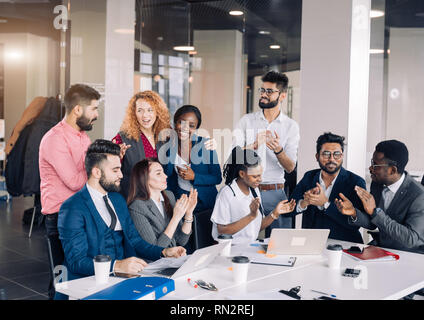 Mixed-Race Gruppe von neun verschiedenen glückliche Menschen stehen rund um die Tabelle mit den Kollegen sitzen, applaudieren Team Leistungen Stockfoto