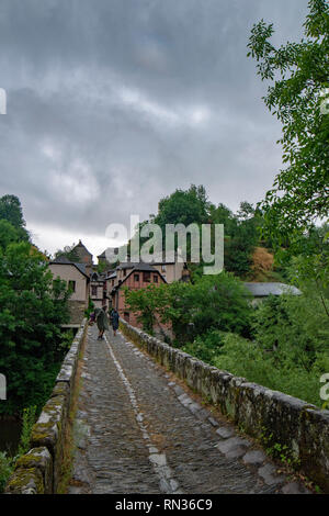 Conques, Frankreich - Juni 2015: eine Gruppe von Pilgern überquert einen kleinen Fluss auf einem Stein Brücke entlang der Hl. Jakobus Pilgerweg in Conques Stockfoto