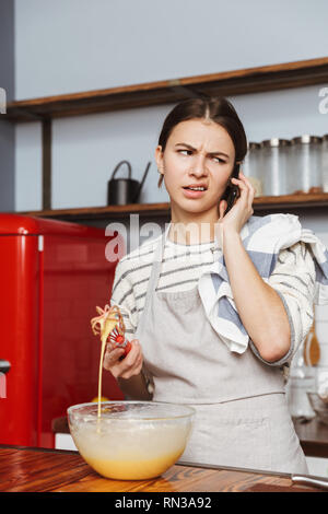 Verwirrte junge Frau steht in der Küche zu Hause, Eier in einer Schüssel mischen, Gespräch am Handy Stockfoto