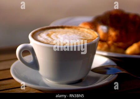 Köstlich heißen italienischen Cappuccino steht in eine Tasse mit Untertasse auf einem Holztisch. Im Hintergrund ein frisches Croissant auf einem Teller. Stockfoto