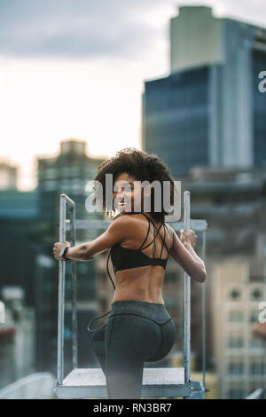 Afro-amerikanerin in Fitnesskleidung klettert die Dachtreppe runter. Fitness-Frau, die auf dem Dach steht und zurückblickt. Stockfoto