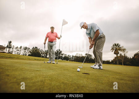 Männliche Golf Spieler einen Schuß auf Putting Green mit einem zweiten Spieler im Hintergrund halten die Flagge. Professionelle Golfspieler zusammen Golf spielen auf t Stockfoto