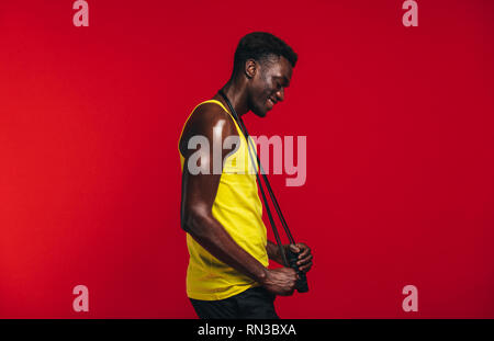 Seitenansicht der Afrikanischen muskulösen Mann mit dem springen Seil um den Hals auf rotem Hintergrund. Lächelnd männlich fitness Modell mit Springseil. Stockfoto