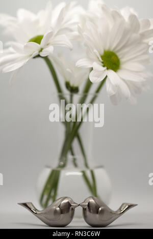 Zwei silberne Liebe vogel Figuren vor der Gänseblümchen in Glas bud vase Küssen auf grauem Hintergrund Stockfoto