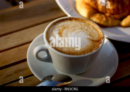 Köstlich heißen italienischen Cappuccino steht in eine Tasse mit Untertasse auf einem Holztisch. Im Hintergrund ein frisches Croissant auf einem Teller. Stockfoto