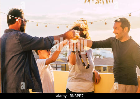 Gruppe von Freunden Toasten und Anstoßen mit Bier und Orangensaft zusammen in den Sonnenuntergang auf der Dachterrasse mit Blick auf die Stadt - gemischten Alters ein Stockfoto