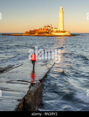 Ein Mann steht auf der Hl. Maria Island Causeway, während die untergehende Sonne St Mary's Leuchtturm an der Küste von Whitley Bay leuchtet in Tyneside. Stockfoto